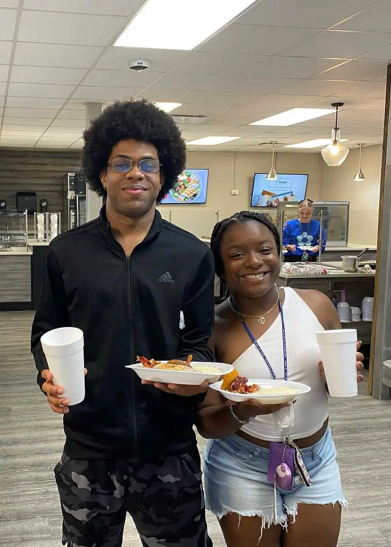 Two students holding plates of food and smiling while standing in cafeteria